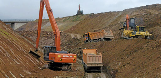 Der Lastwagen in der Mitte fährt auf dem Niveau der künftigen Umgehung, links und rechts fahren die Lastwagen und arbeiten die Bagger auf einem aufgeschüttenen Damm, der die notwendige Arbeitshöhe bringt.